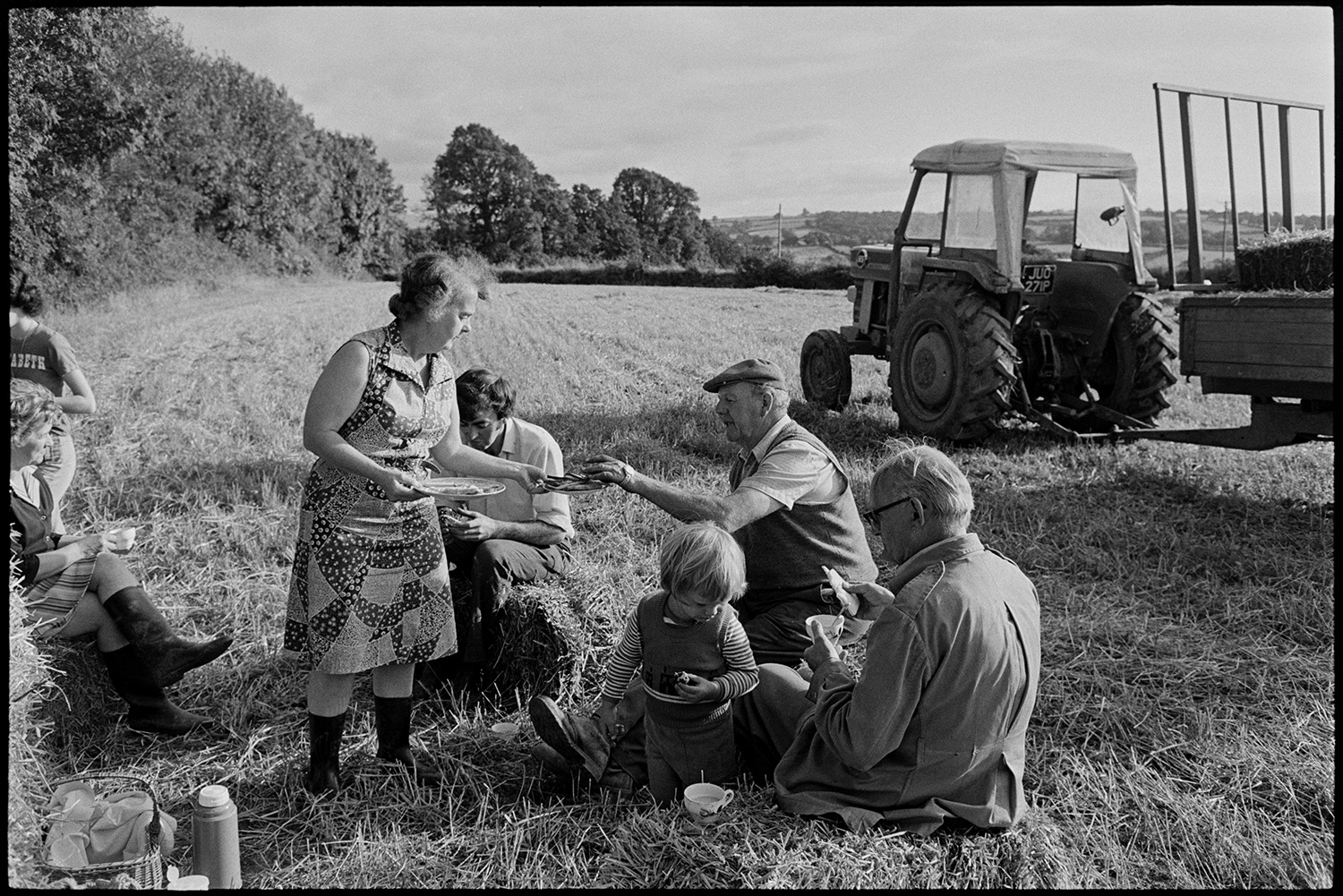 Harvesters tea in field. 
[The Ward family having a tea break from harvesting in a field at Nethercott, Iddesleigh. Hettie Ward is handing out food and a tractor and trailer is parked in the background.]