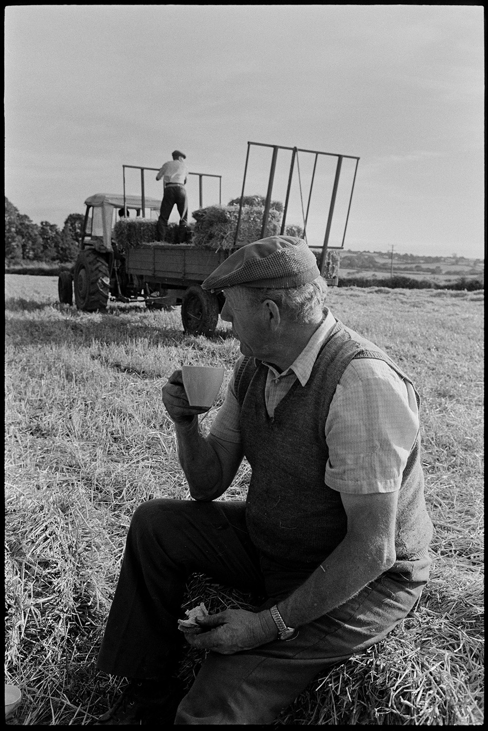 Harvesters tea in field. 
[John Ward sat on a bale of straw and drinking a cup of tea in a field at Nethercott, Iddesleigh, while he has a break from harvesting. A man is stood with straw bales on a trailer in the background.]