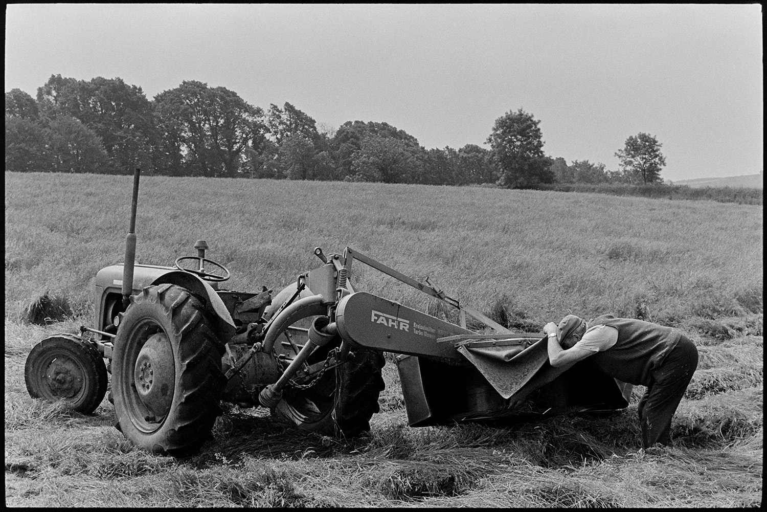 Grass cutter and tractor, farmer checking. 
[John Ward checking a grass cutter and tractor in a field at Parsonage, Iddesleigh.]