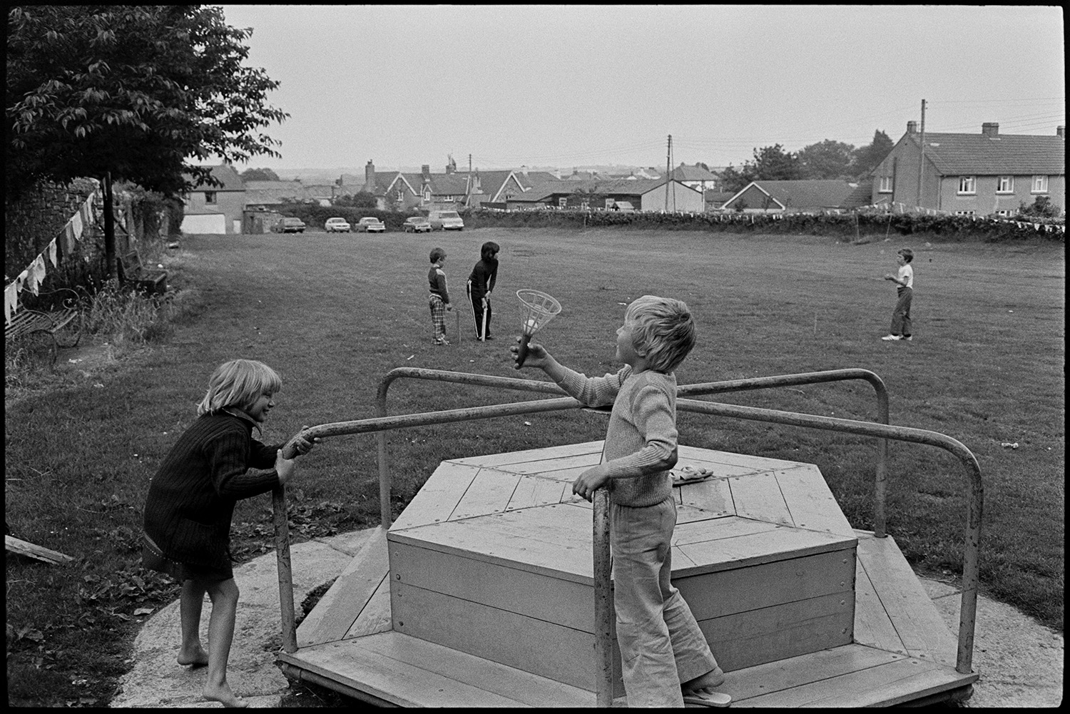 Village playground, children. 
[Children playing on a roundabout in the village playing field at Beaford. One of the children is holding a cup and ball. More children can be seen playing cricket behind them. In the background houses and parked cars are visible.]