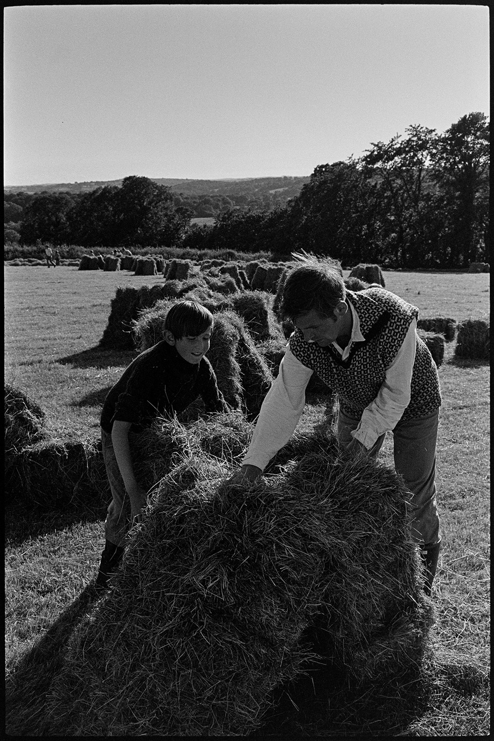 Children watching and helping farmer bale hay. 
[A boy helping Michael Morpurgo stack hay bales together in a field at Parsonage, Iddesleigh.]