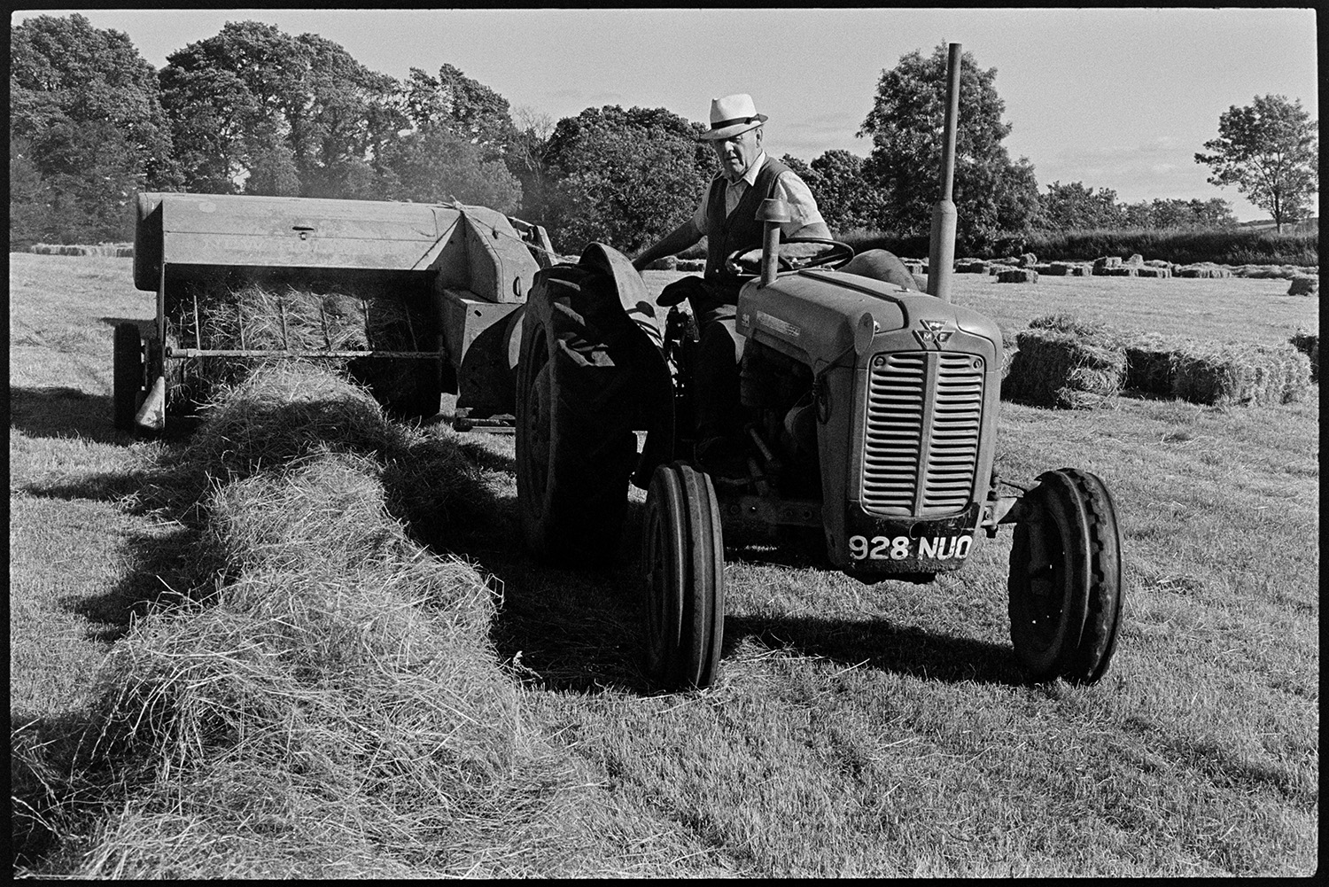 Children watching and helping farmer bale hay. 
[John Ward baling hay with a tractor and baler in a field at Parsonage, Iddesleigh. Other hay bales can be seen in the field in the background.]