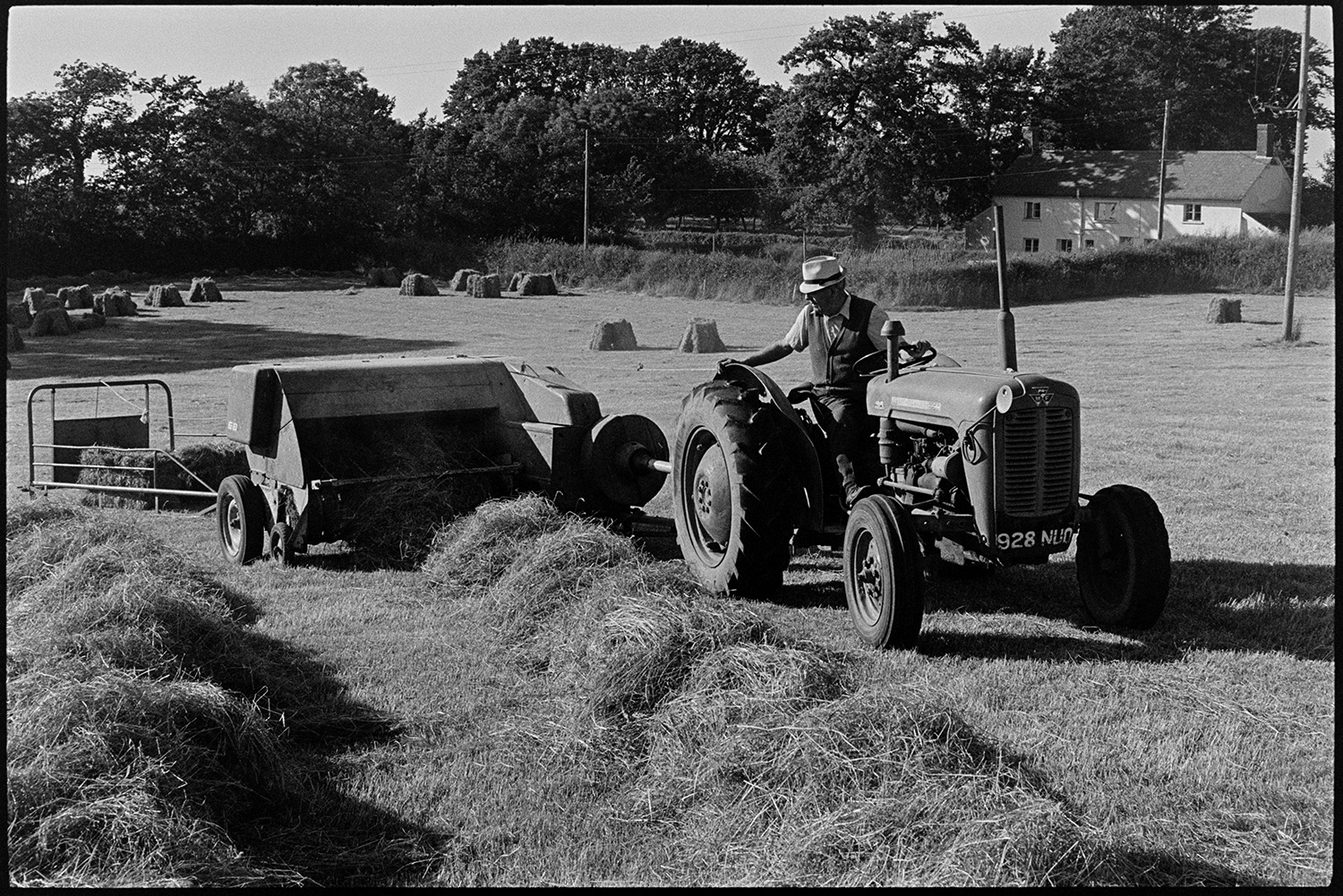 Children watching and helping farmer bale hay. 
[John Ward baling hay with a tractor and baler in a field at Parsonage, Iddesleigh. Stacks of hay bales can be seen in the field in the background.]