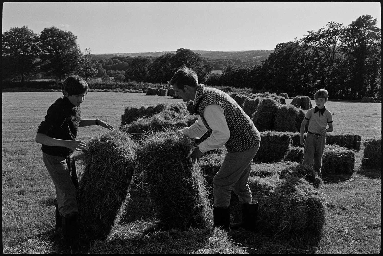 Children watching and helping farmer bale hay. 
[Two boys helping Michael Morpurgo stack bales of hay in a field at Parsonage, Iddesleigh.]