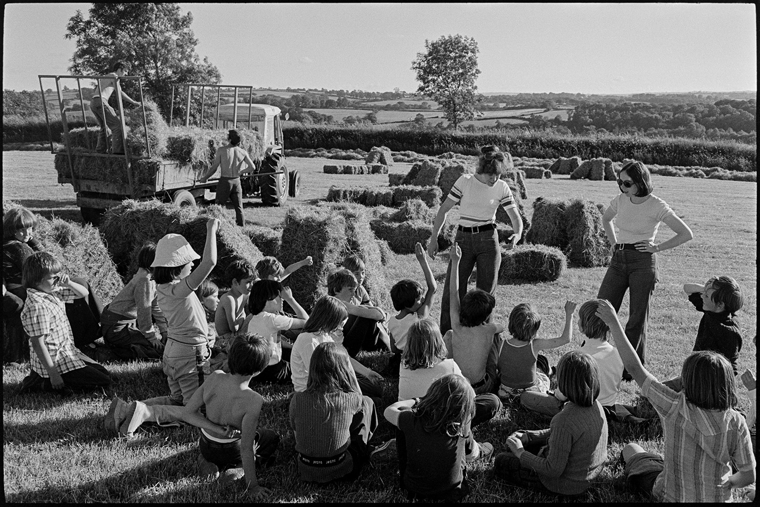 Children watching and helping farmer bale hay. 
[Children sat in a field at Parsonage, Iddesleigh, answering questions from their teachers at the Farms for City Children initiative. In the background two men are loading hay bales onto a tractor and trailer.]