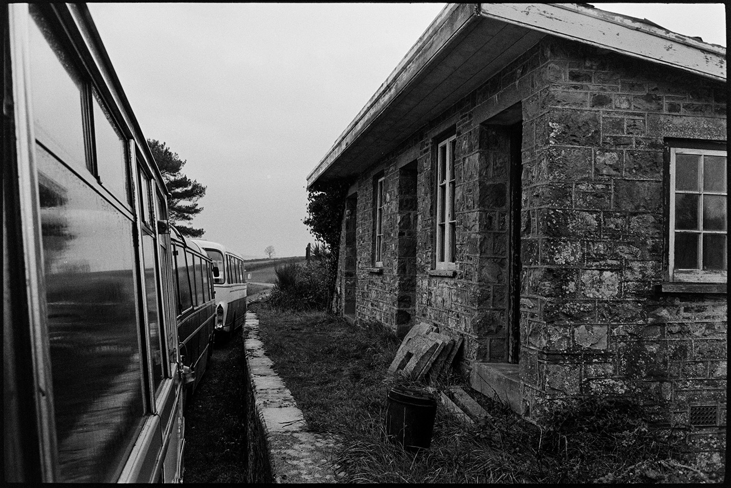 Disused railway station with parked buses. 
[The disused building of Meeth Railway Station with three buses parked outside. The platform is overgrown with grass and brambles.]