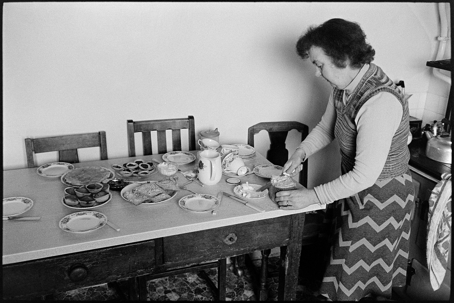 Woman, farmers wife cooking and making tea. 
[Hettie Ward preparing tea in her kitchen at Parsonage, Iddesleigh. The kitchen table is laid with cakes, plates and teacups. Hettie is cutting slices of bread. A kettle can be seen on a rayburn in the background.]