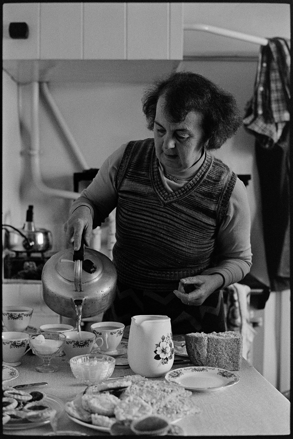 Woman, farmers wife cooking and making tea. 
[Hettie Ward making cups of tea in her kitchen at Parsonage, Iddesleigh. She is pouring hot water in to the teacups from a kettle. The kitchen table is laid for tea with cakes and slices of bread.]