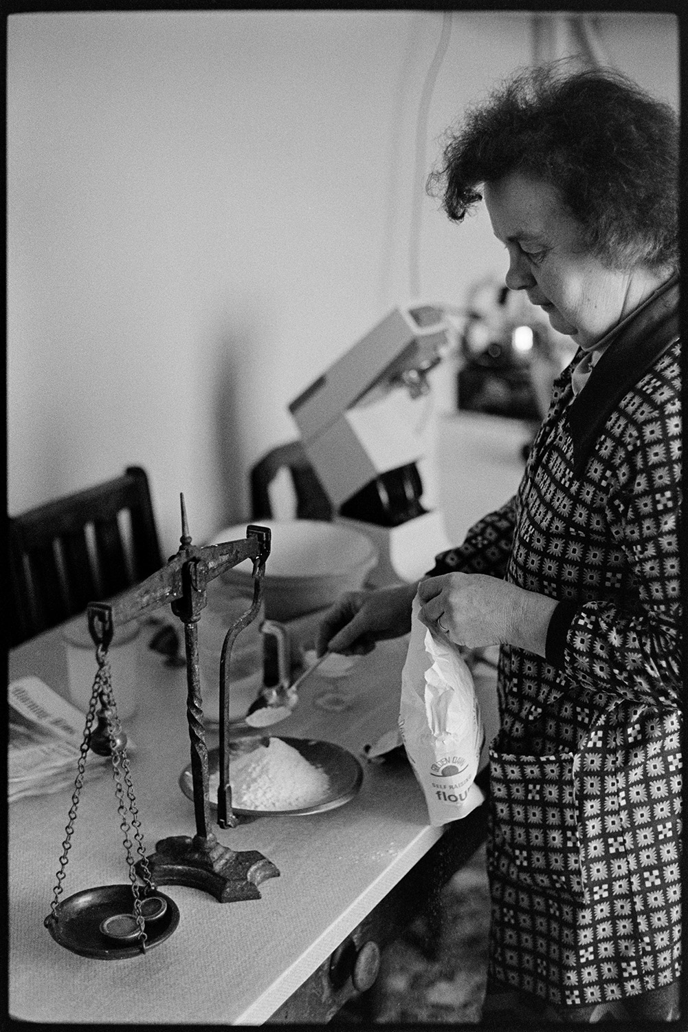 Woman weighing flour. 
[Hettie ward weighing flour using a set of scales with weights, in her kitchen at Parsonage, Iddesleigh.]
