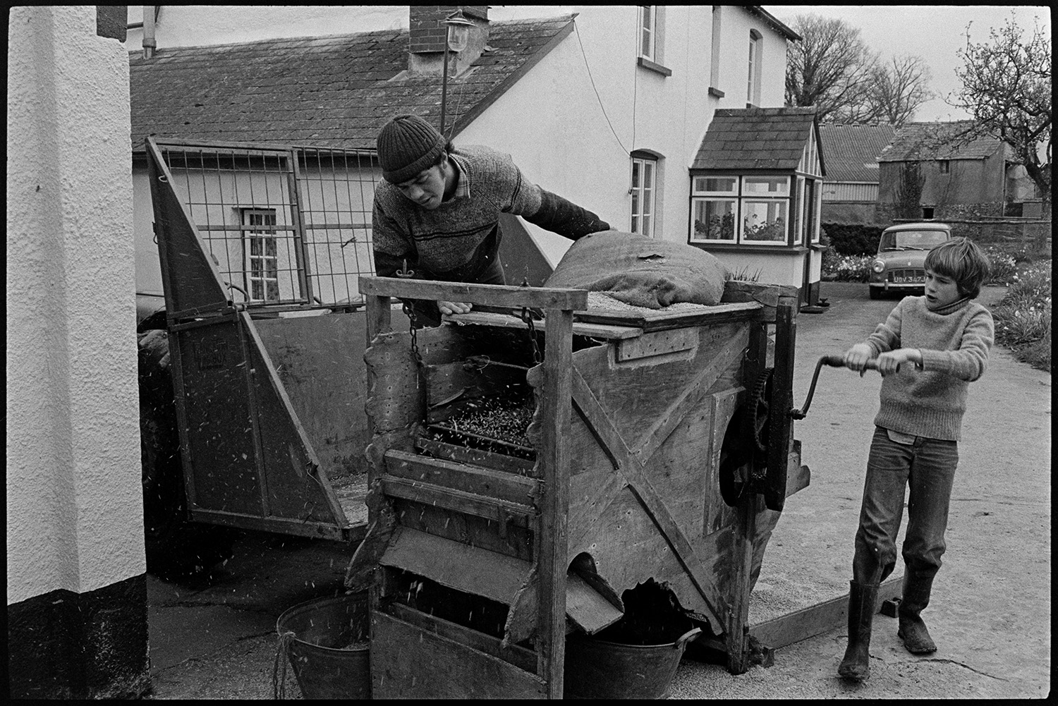 Men operating old crusher, hand operated. 
[David or Graham Ward and a boy using a grain crusher at Parsonage, Iddesleigh. The boy is turning the handle to work the crusher while the other man is checking the crusher. The farmhouse and a parked car can be seen in the background.]