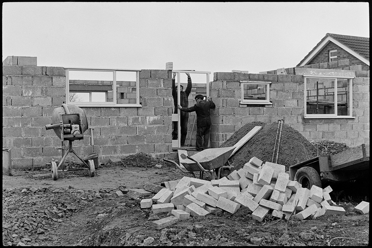 Bungalow, house being built. 
[Two men building a bungalow in Dolton, They are putting in the door frame. A cement mixer, wheelbarrow, pile of earth and bricks can be seen in the foreground.]