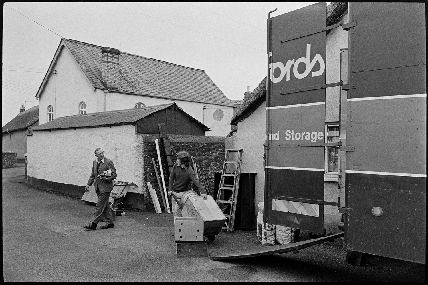 Removal van outside house. 
[A man loading a box into a removal van in West Lane, Dolton. He is talking to a man passing by. A ladder, sacks and shelves are visible on the roadside.]