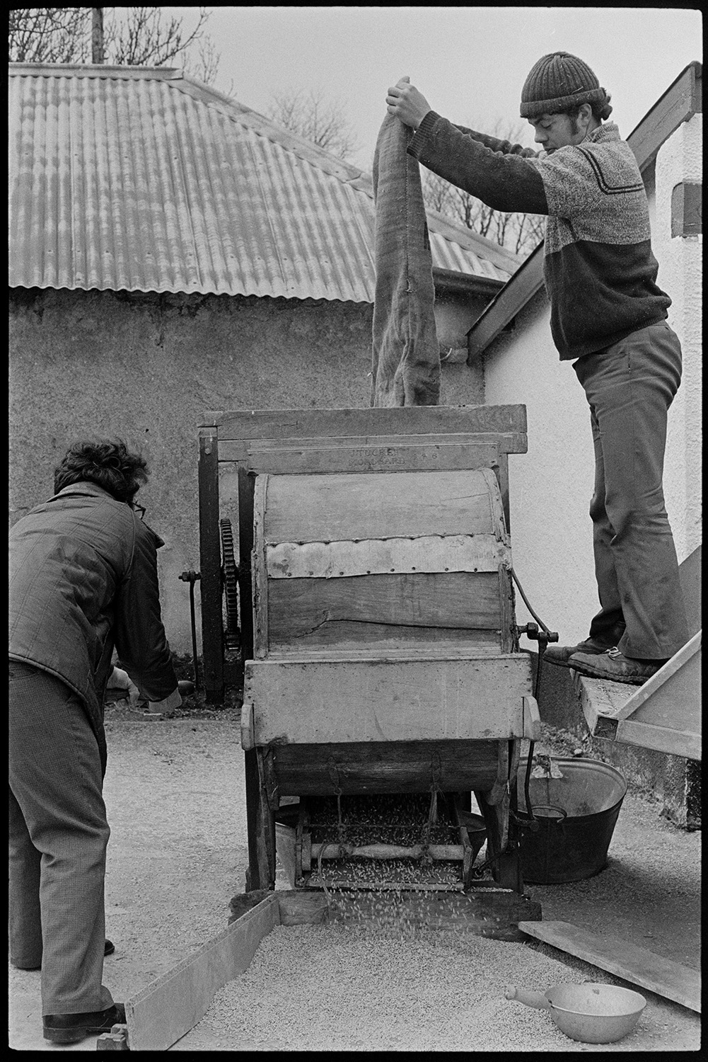Men operating old crusher, hand operated. 
[David or Graham Ward and another man using a grain crusher in the farmyard at Parsonage, Iddesleigh. One man is emptying a sack of grain into the crusher and the other man is operating the crusher by turning a handle.]