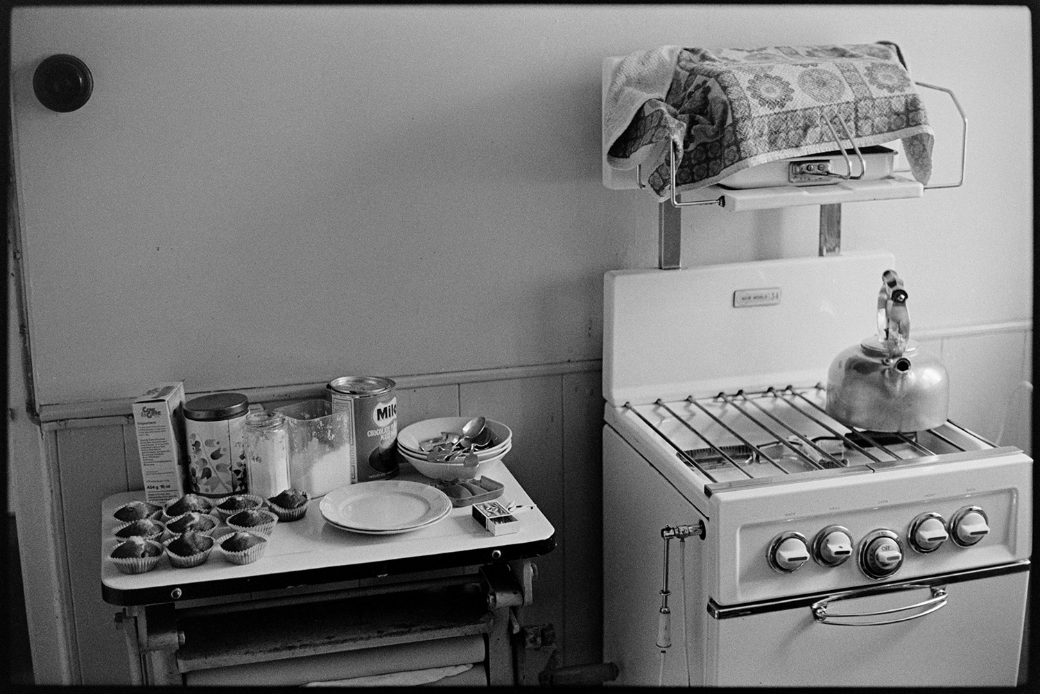 Gas cooker, kettle with buns on sideboard. 
[A kettle on a gas cooker in a house, possible Emily Easterbrook's, in West Lane, Dolton. A side table with plates, bowls, spoons and cakes is next to the cooker.]