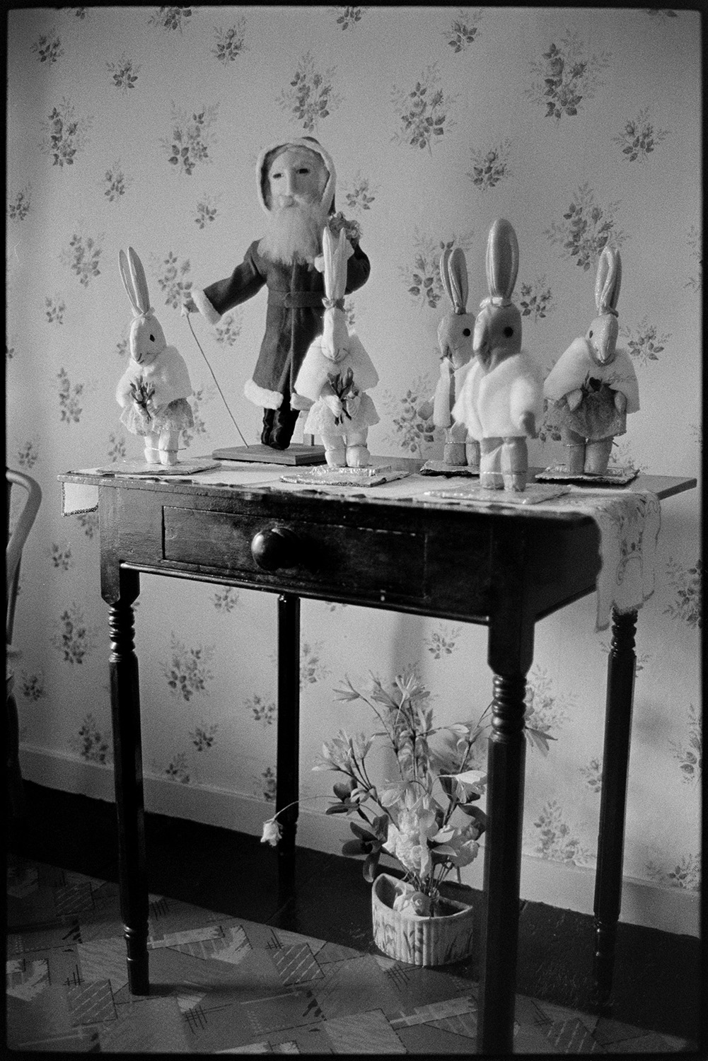 Stuffed toys on table, hand made. 
[Emily Easterbrook's hand made toys, including a Father Christmas and rabbits, displayed on a side table in her home at West Lane, Dolton. The wall is covered in floral patterned wallpaper and a flower display is underneath the table.]