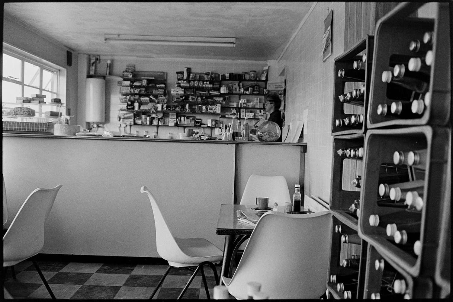 Interior of cafe at garage, sweets and soft drinks on shelf. 
[A woman stood behind the counter in a café at the Fishleigh Rock Garage in Umberleigh. Tables and chairs can be seen next to shelves with bottles.]