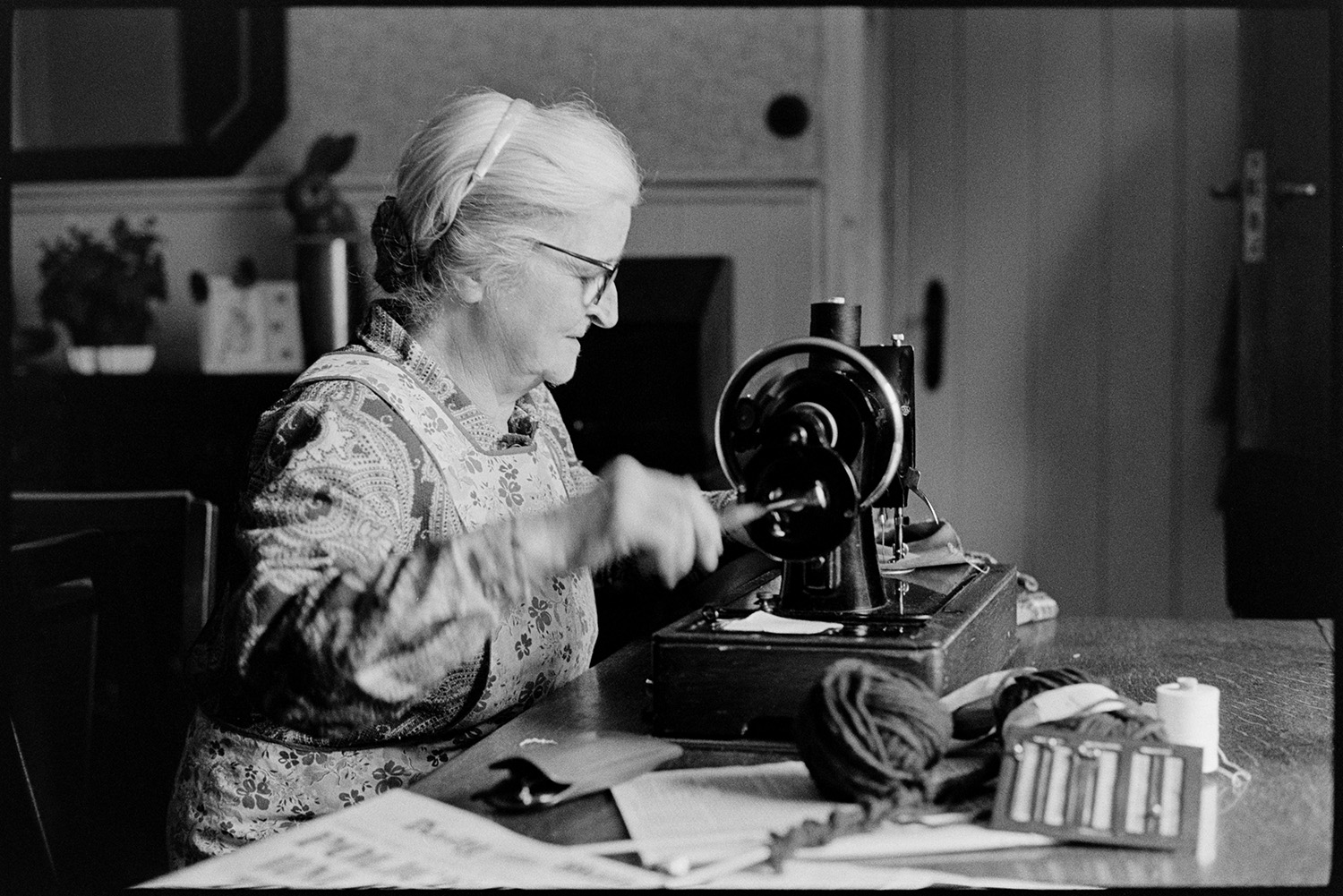 Woman using sewing machine. 
[Norah Maynard using a manual sewing machine in her home at Atherington. A glasses case and ball of wool or thread is on the table.]
