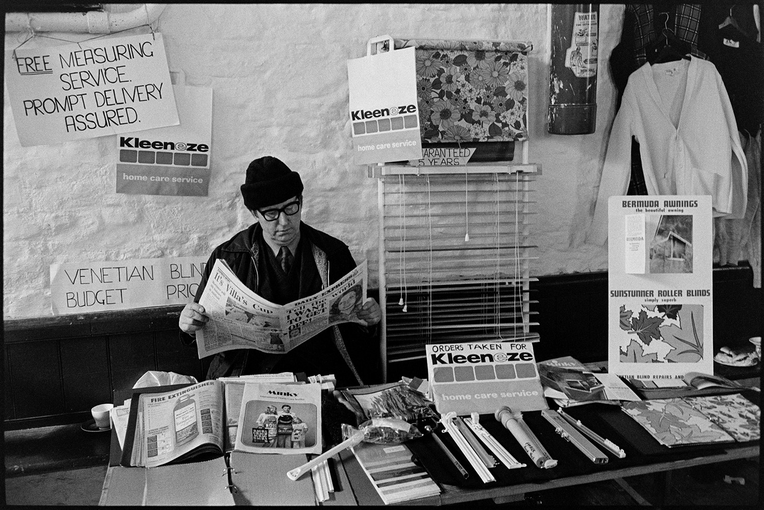 Pannier market, venetian blind stall, Kleeneze products. 
[A man running a stall selling venetian blinds and Kleeneze items at Bideford Pannier Market. He is sat behind the stall reading the Daily Express newspaper.]