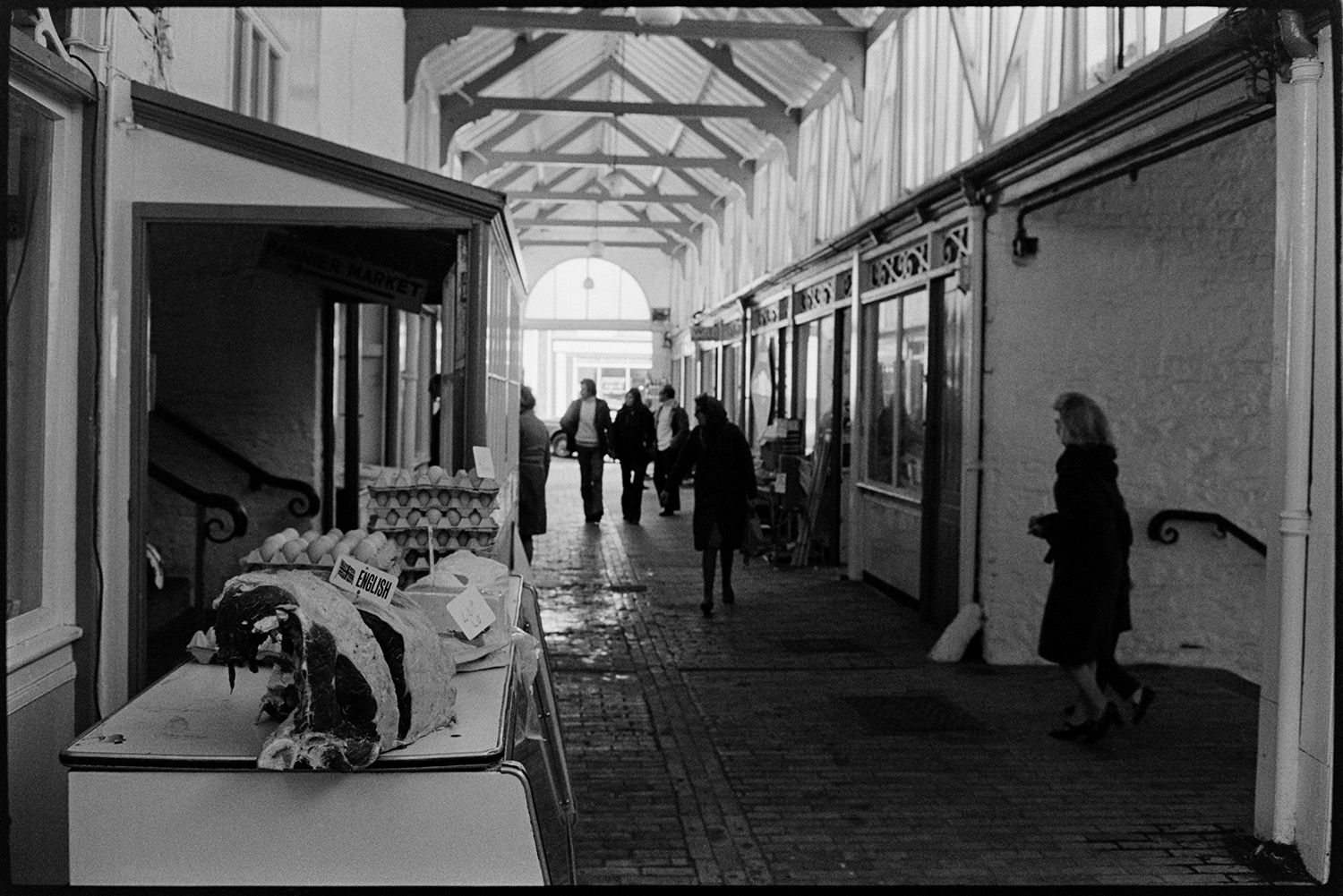 Pannier market, trug ,boots. 
[People walking along a passageway inside Bideford Pannier Market. In the foreground is a stall with cuts of meat and eggs for sale.]