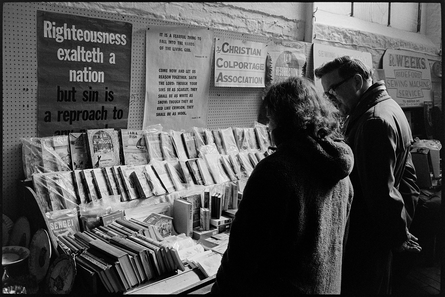 Pannier market, Christian Colportage Association book stall. 
[A man and woman looking at the Christian Colportage Association book stall at Bideford Pannier Market. Religious posters are displayed on the wall behind the book stall.]