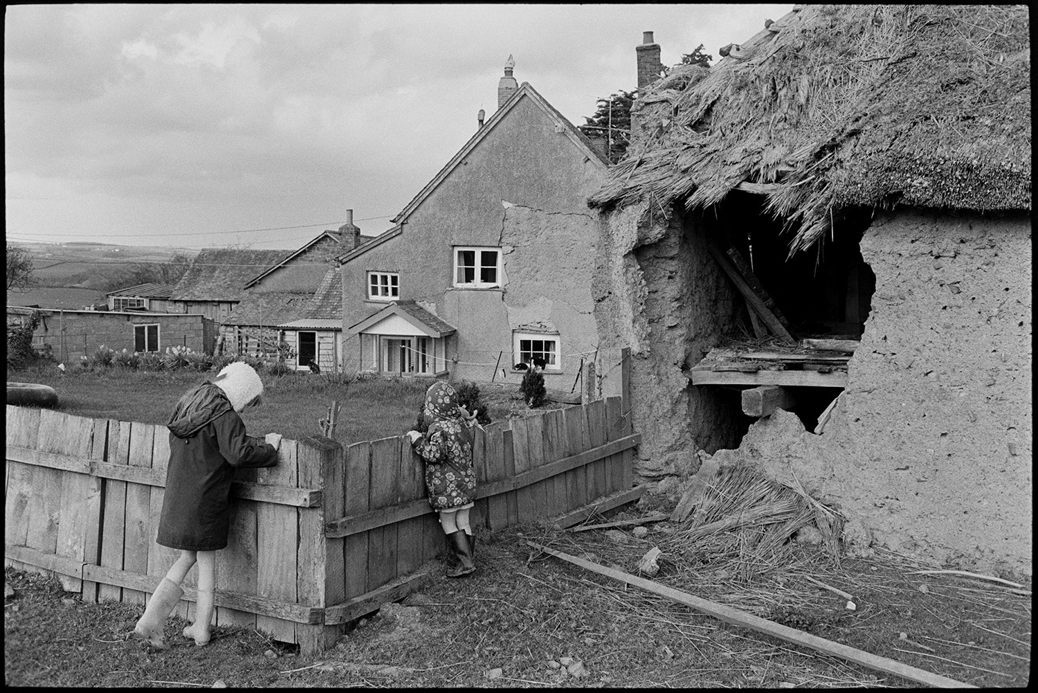 Cob and thatch barn, collapsing. 
[Two children playing by a collapsing cob and thatch barn at Higher House Farm, Atherington. They are looking over a wooden fence to the farmhouse and garden.]