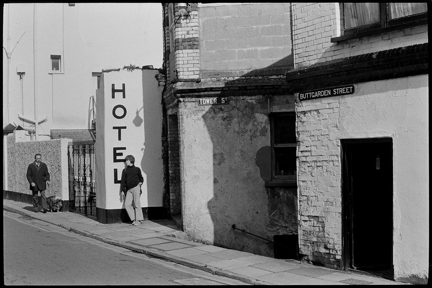 A man walking a dog along Buttgarden Street, Bideford. Another person is leaning against a wall with the word 'Hotel' painted on it.
