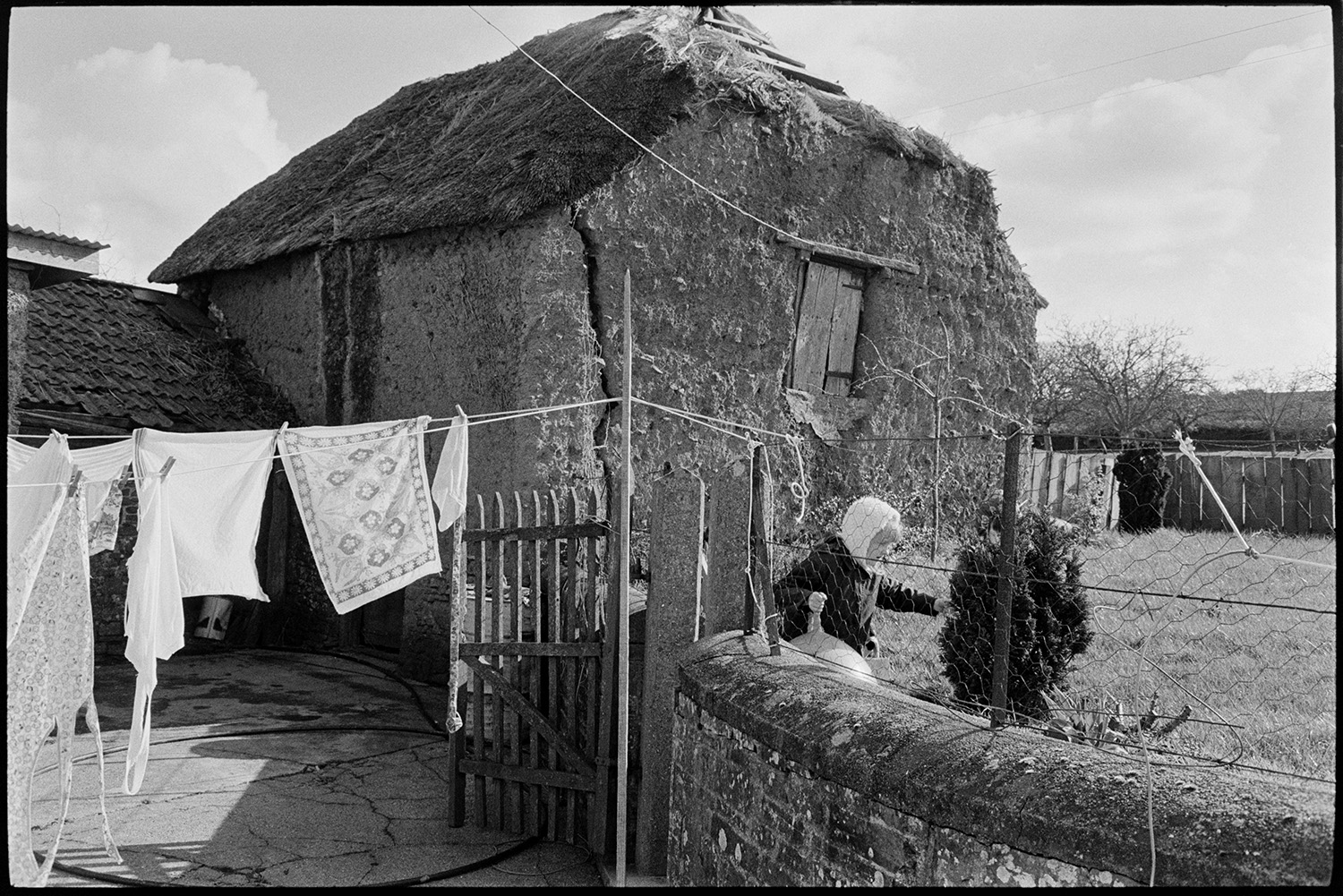 Cob and thatch barn, collapsing. 
[A collapsing cob and thatch barn with a tallet at Higher House Farm, Atherington. The barn wall has a large crack in it. A girl is playing with a space hopper in the garden next to the barn and washing is hanging out to dry on a washing line in the foreground.]