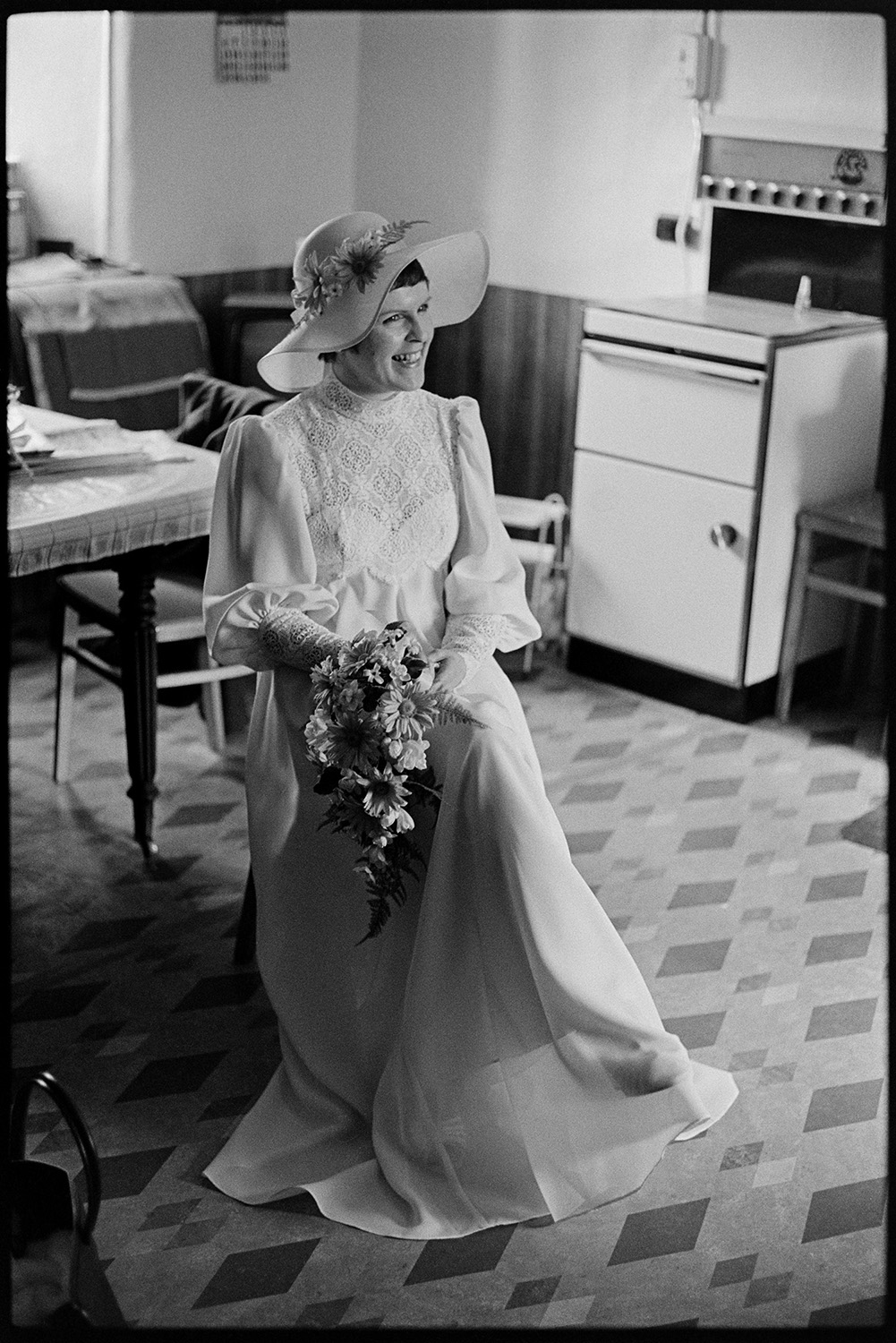 Bride and family at home, setting off for wedding in car with bridesmaid. 
[Mary Pugsley sat on a chair in her kitchen at Lower Langham, Dolton. She is holding a bouquet and wearing a hat decorated with flowers. A cooker can be seen in the background.]