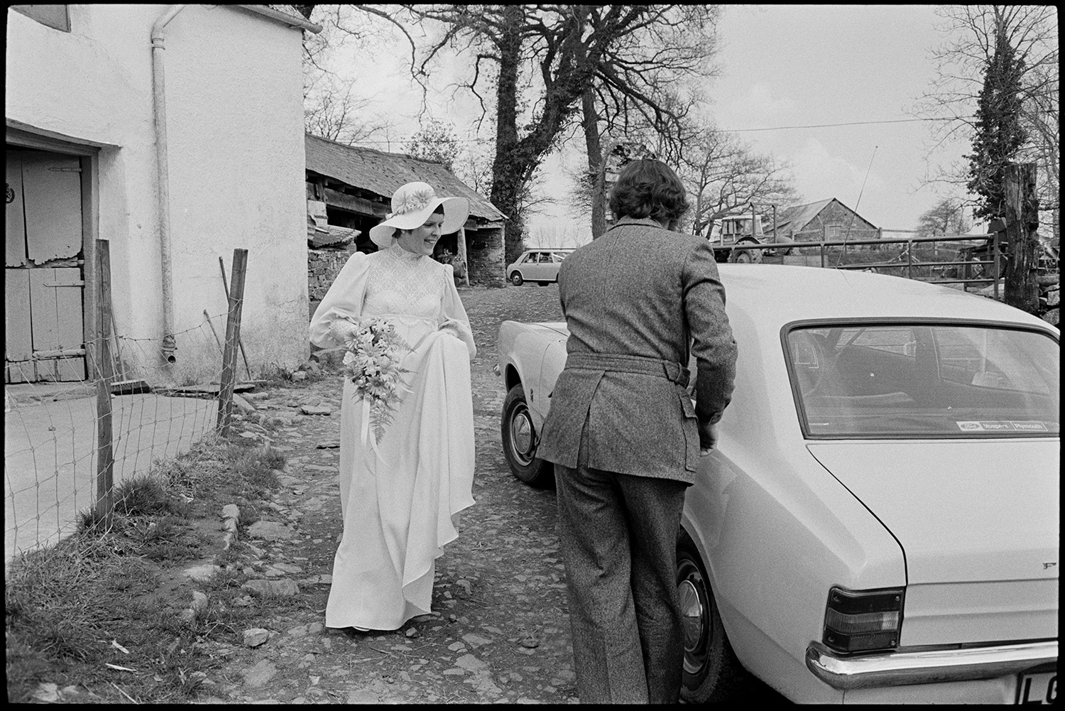 Bride and family at home, setting off for wedding in car with bridesmaid. 
[Mary Pugsley getting into a car in the farmyard at Lower Langham, Dolton, to go to her wedding. She is carrying a bouquet and wearing a hat decorated with flowers. Barns and a tractor can be seen in the background.]