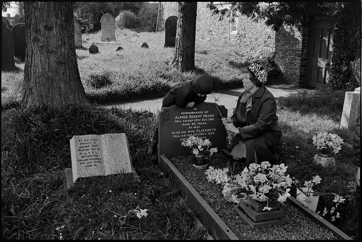 People putting flowers on graves at Easter time. 
[A woman and child putting flowers on the grave of Alfred Heard and Elizabeth Geard in Dolton Churchyard at Easter. The woman is holding a daffodil. Trees and gravestones are visible in the background.]