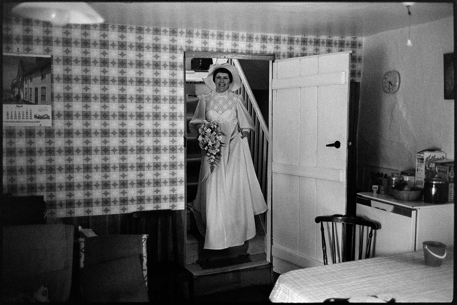 Bride and family at home, setting off for wedding in car with bridesmaid. 
[Mary Pugsley in her wedding dress at the bottom of the stairs in her house at Lower Langham, Dolton, before her wedding. She is holding a bouquet and wearing a hat decorated with flowers. A kitchen table can be seen in the foreground and one of the walls is covered with patterned wallpaper.]