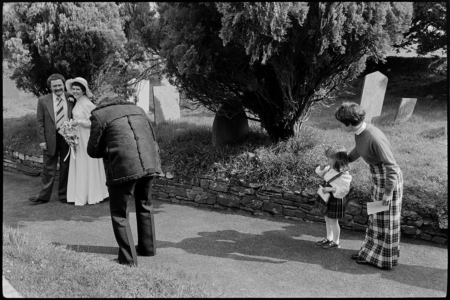 After wedding, bride, groom and guests, photographer at work. 
[A photographer taking pictures of Mary Pugsley and her groom after their wedding, in Dolton Churchyard. Two wedding guests, a woman and young girl, are watching.]