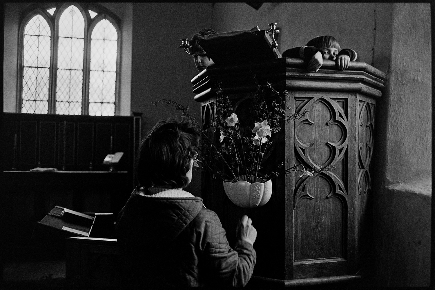 People putting Easter flowers in Church. 
[Mrs Folland arranging a flower display for Easter on the pulpit in Dowland Church. A child is peeping over the top of the pulpit.]