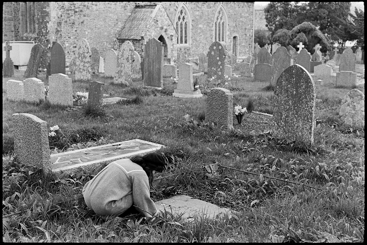 People tending graves. 
[Robin Ravilious putting flowers on a grave in Dolton Churchyard. The church can be seen in the background behind other gravestones.]