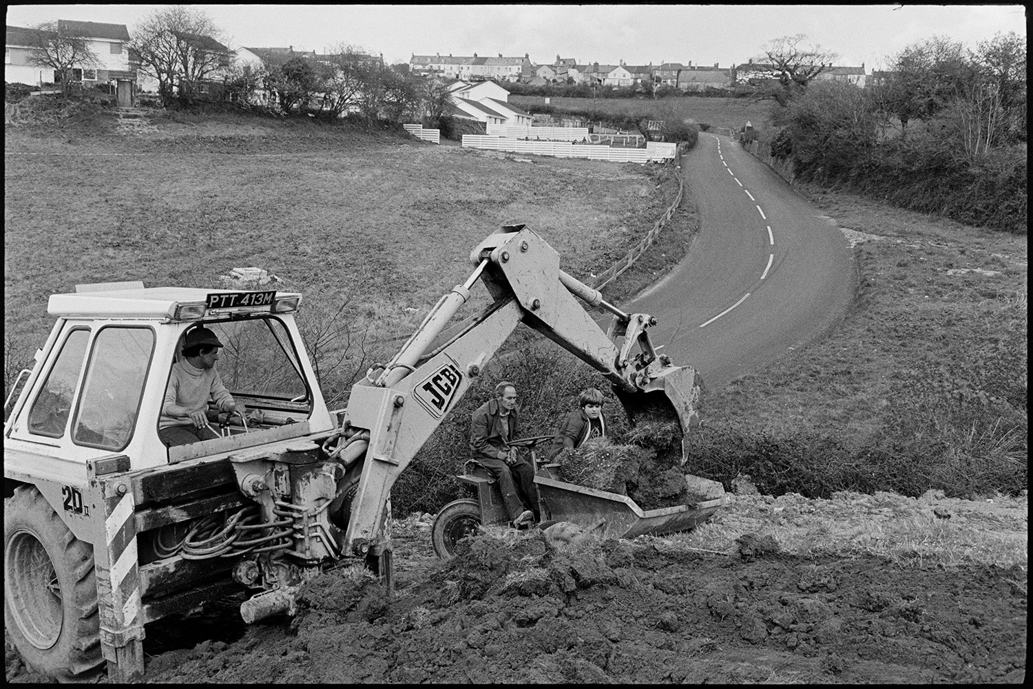 JCB and dumper truck excavating building site. 
[A JCB digger stripping topsoil from a building site in Torrington and placing it into the bucket of a dumper truck. A man and boy are sat behind the controls of the dumper truck. A road and houses can be seen in the background.]