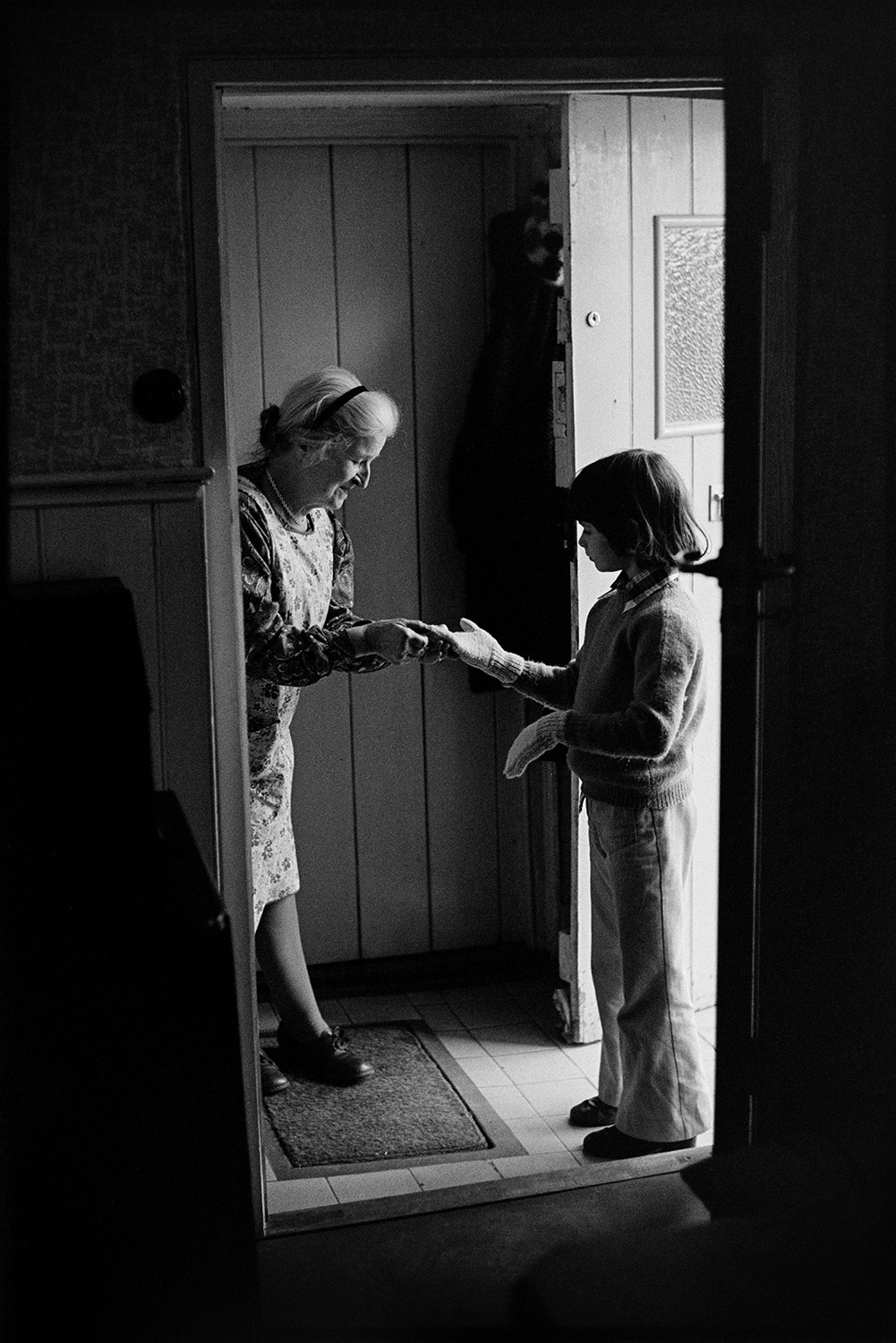Woman talking to child at door. 
[Norah Maynard talking to a child at the front door of her house, named White Hart, in Atherington. She is helping the child put on a glove.]