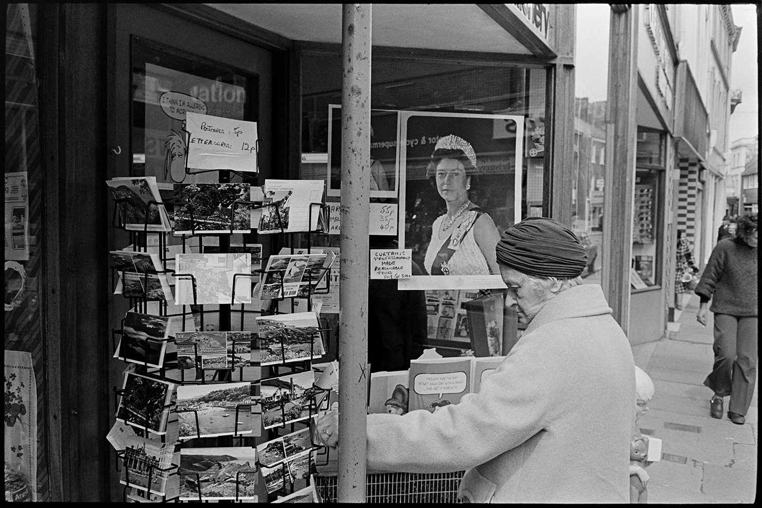 A woman looking at postcards on display outside a newsagents in Barnstaple. A picture of Queen Elizabeth II is displayed in the shop window.