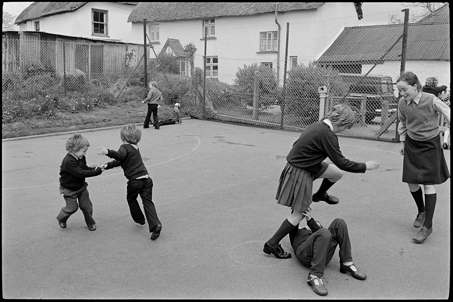 In and out of school with children playing. 
[Schoolchildren playing in the playground at Kings Nympton Primary School. A thatched cottage is visible behind the school fence.]