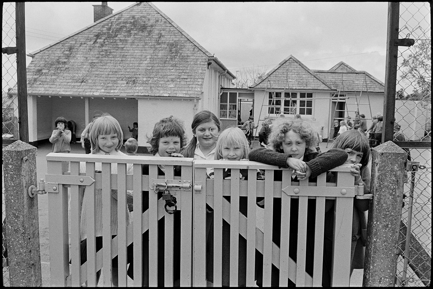 In and out of school with children playing. 
[Schoolchildren looking over the school gate at Kings Nympton Primary School. Children can be seen playing on a climbing frame in the playground in the background.]