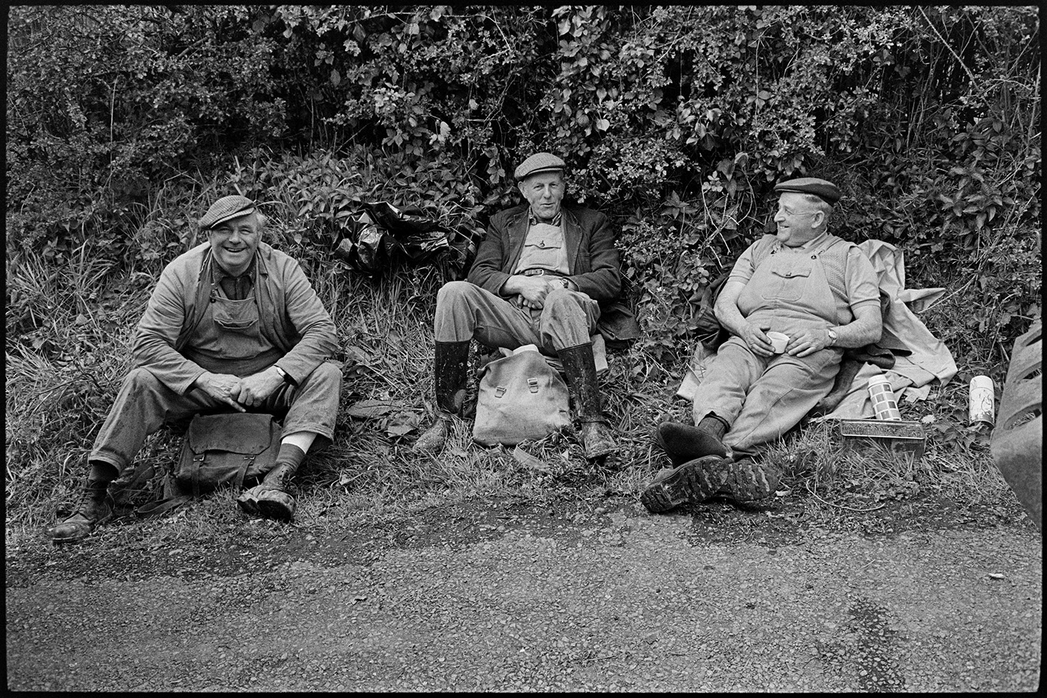Road menders having lunch, see pic in old archive at same spot. 
[Three men sat on a roadside verge at Kings Nympton, eating their lunch. They have been doing repairs to the road.]