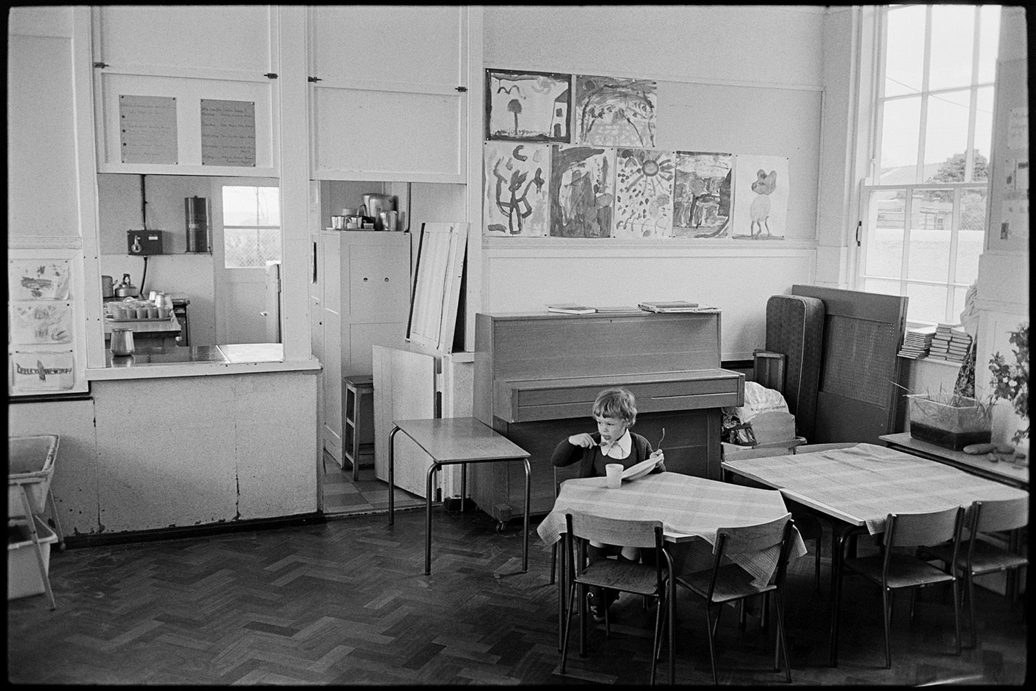 In and out of school with children playing. 
[A child eating at a table in the hall at Kings Nympton Primary School. A piano and serving hatch to the school kitchen can be seen in the background. Children's artwork is displayed on the wall.]