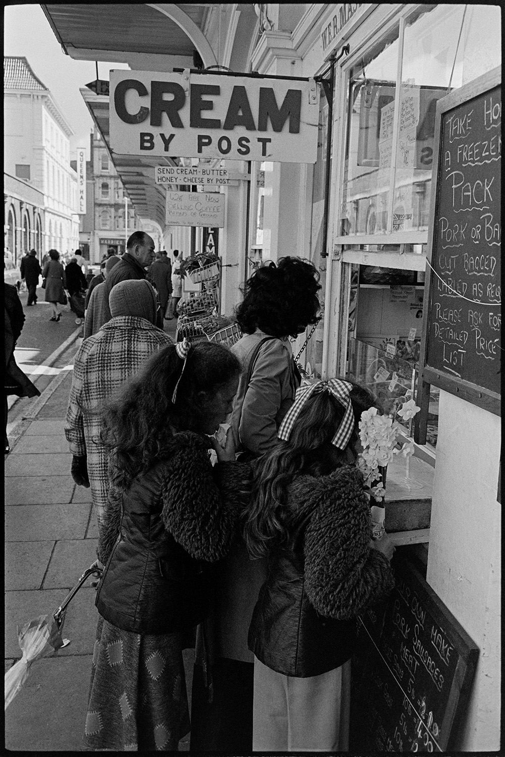Row of shops with shoppers, displays of flowers and fruit. 
[A woman and two girls buying meat from a butcher's shop hatch in Butcher's Row, Barnstaple. A sing hanging above their heads reads 'Cream By Post'.]