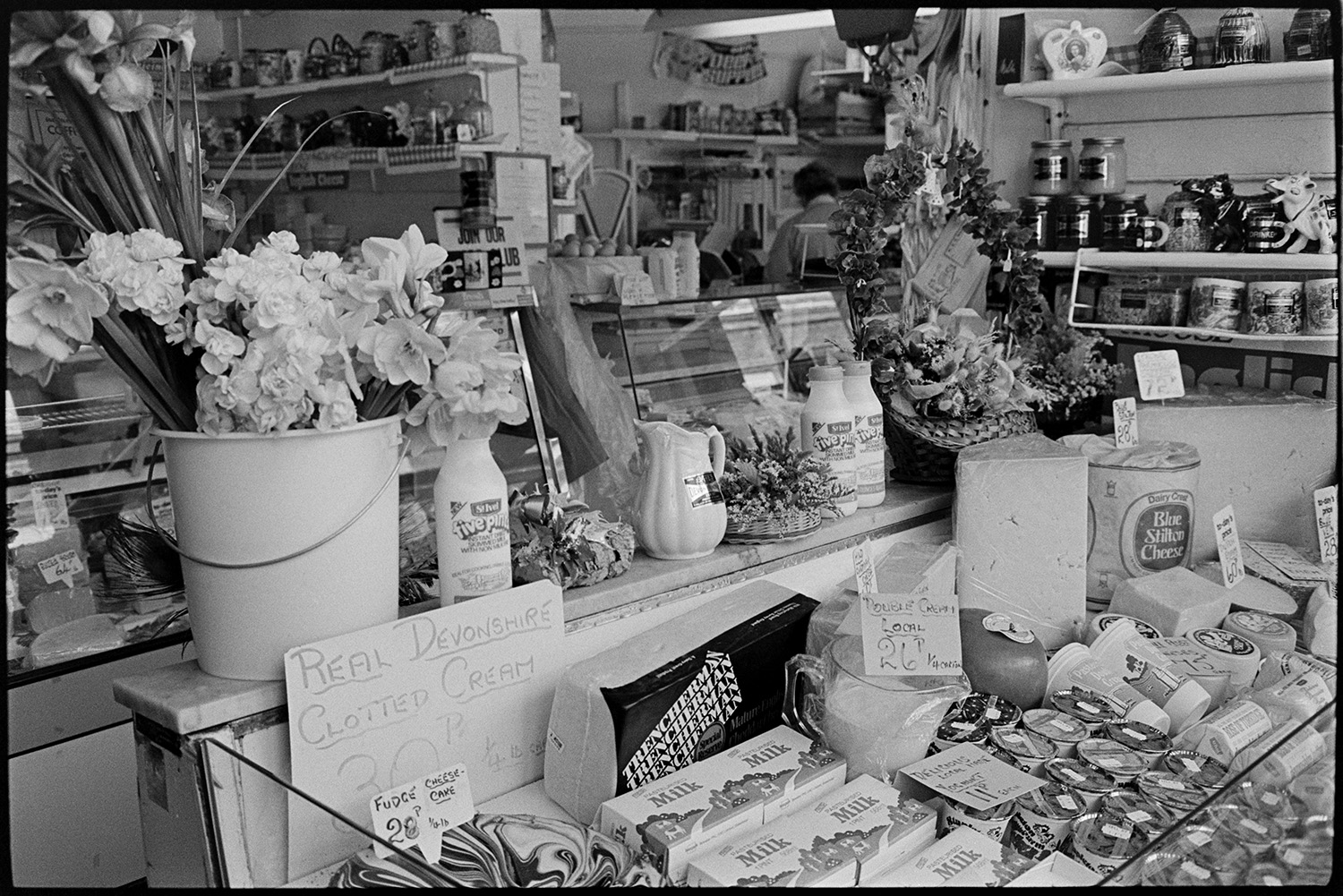 Row of shops with shoppers, displays of flowers and fruit. 
[A refrigerated display of cheese, cartons of milk, yoghurt and cream in a shop in Butcher's Row, Barnstaple. A bucket of daffodils, and jars of honey can be seen on the shelves in the shop, amongst other goods.]