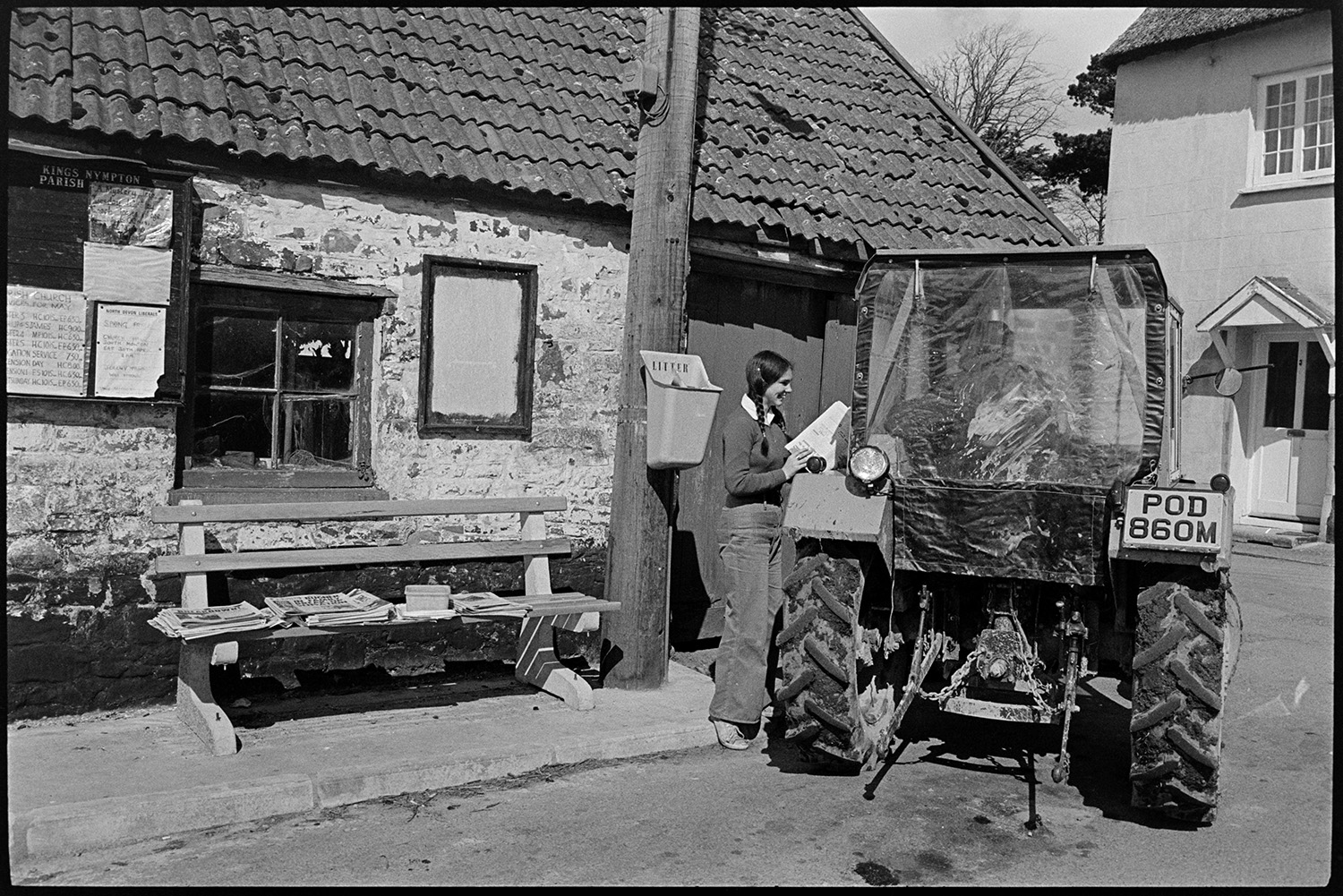 Sunday papers being sold in village, farmer in tractor and people chatting. 
[A girl selling Sunday newspapers in Kings Nympton to a person in a tractor. The newspapers are laid out on a bench. A noticeboard is attached to a building in the background.]