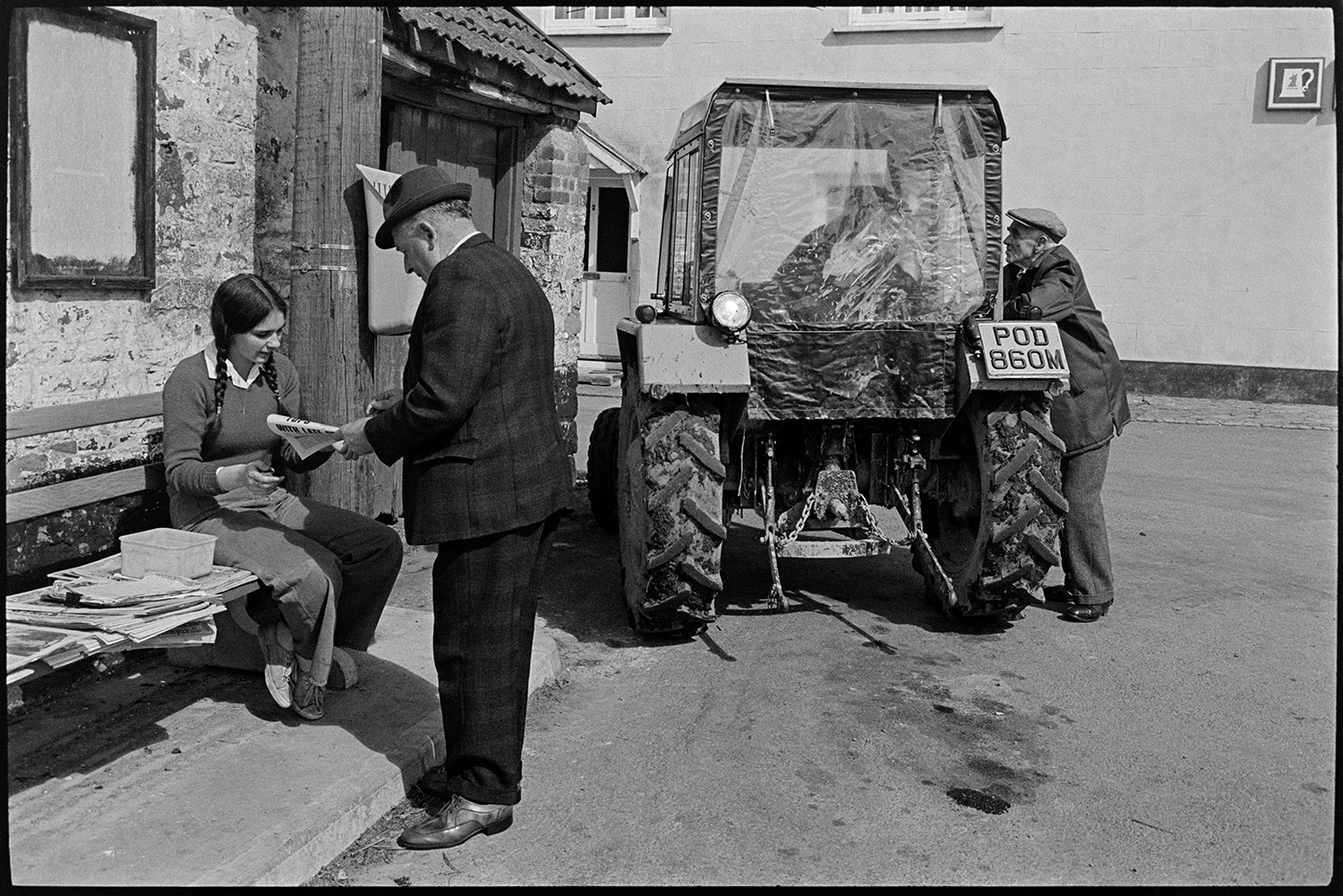 Sunday papers being sold in village, farmer in tractor and people chatting. 
[A girl selling a Sunday newspaper to a man in Kings Nympton. She is sat on a bench with the newspapers. In the background another man is talking to a person in a tractor.]
