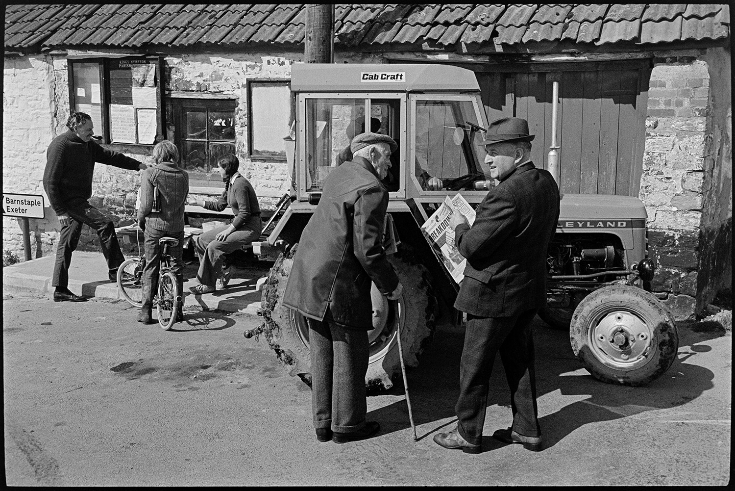 Sunday papers being sold in village, farmer in tractor and people chatting. 
[A girl sat on a bench in Kings Nympton and selling Sunday newspapers to a man smoking a pipe and a person on a bicycle. In the foreground two men are talking to a person in a tractor. One of the men has a walking stick and the other is holding a newspaper which he has just bought.]