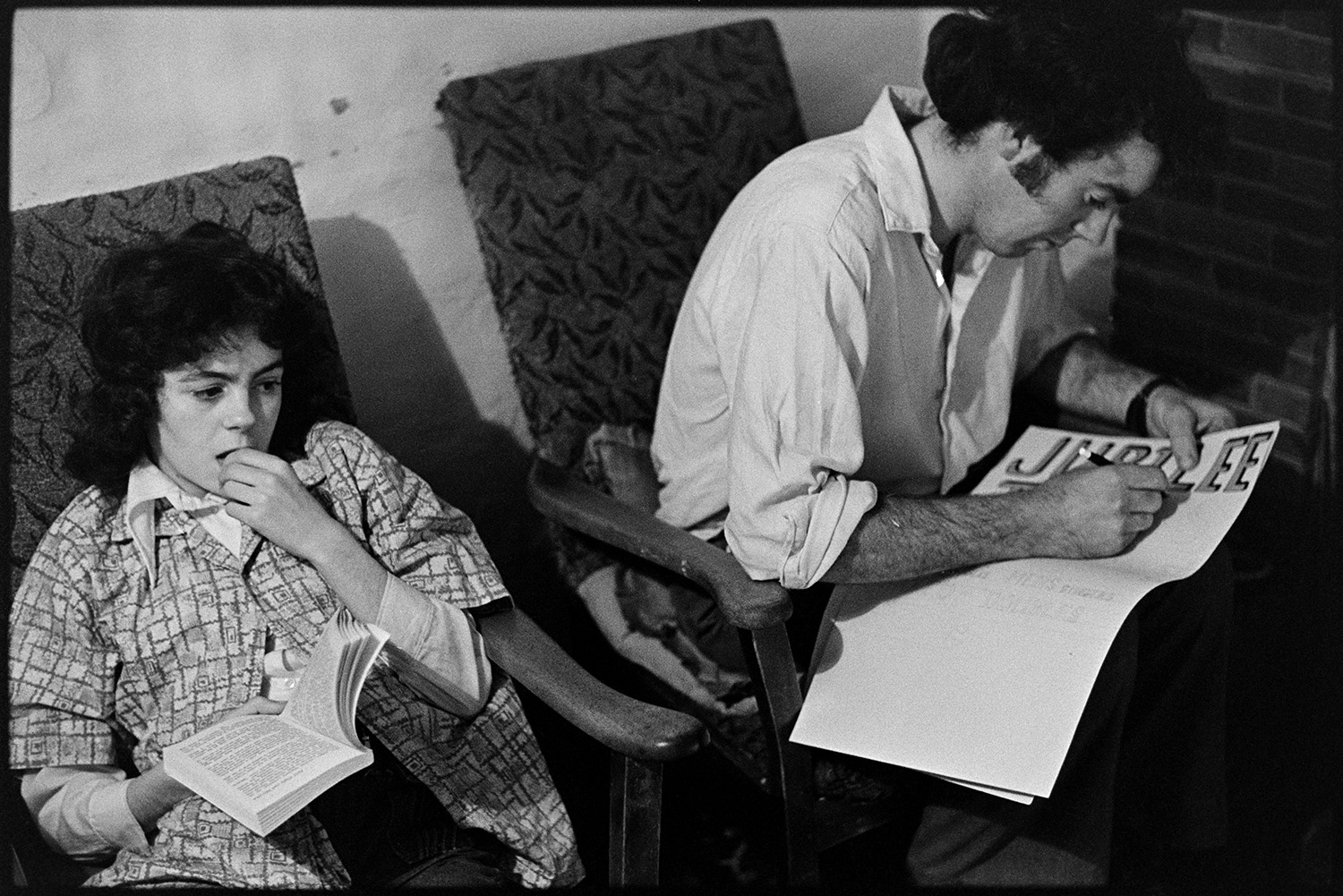 Brother and sister sitting down reading and designing poster. 
[Elizabeth Ward and Graham Ward sat down in their farmhouse at Parsonage, Iddesleigh. Elizabeth is reading a book and Graham is designing a poster for a Jubilee, possibly Queen Elizabeth II Silver Jubilee.]