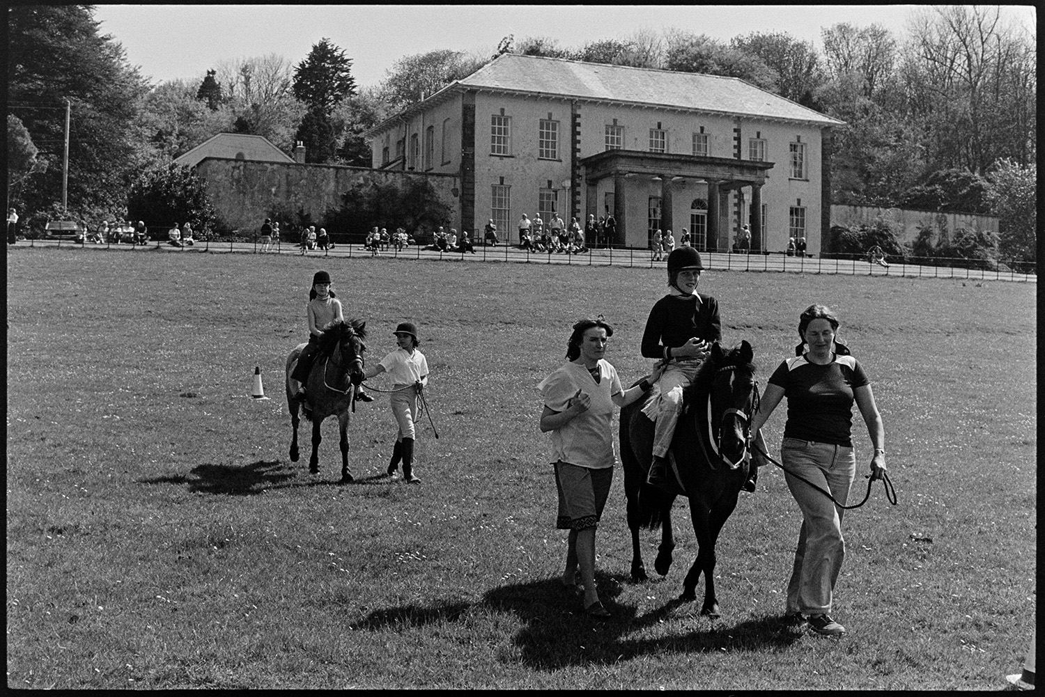 Horse riders and Wombles, Gymkhana, people chatting and having fun in aid of the handicapped. 
[Two children having pony rides at a fete at Cross House, Torrington to raise money for the Riding for the Disabled Association. People are watching the pony rides from a terrace outside the house.]