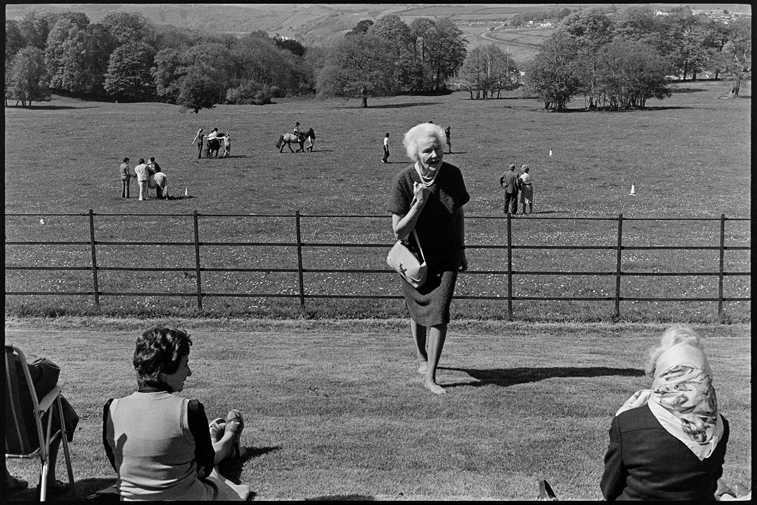 Horse riders and Wombles, Gymkhana, people chatting and having fun in aid of the handicapped. 
[Women sat on a grassy slope outside Cross House, Torrington watching children having pony rides at a fete to raise money for the Riding for the Disabled Association. Lady Fisher is walking towards the women sitting down. One woman is wearing a headscarf. Parkland can be seen in the background.]
