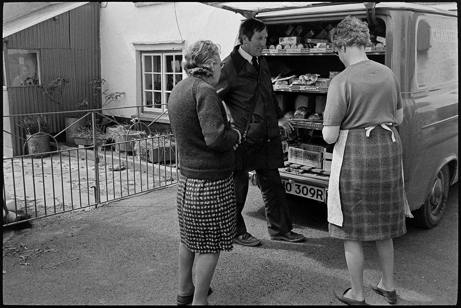 Women buying food from mobile grocers van. <br />
[Miss Audrey Bowden, on the left, and another woman buying groceries from a Grocer's van in a street in Kings Nympton.]